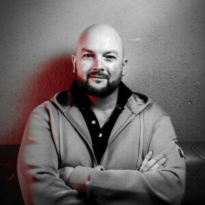 Črno-bela fotografija Neila Bracewella. Nosi siv pulover s kapuco in stoji s prekrižanimi rokami ter se smehlja v kamero.